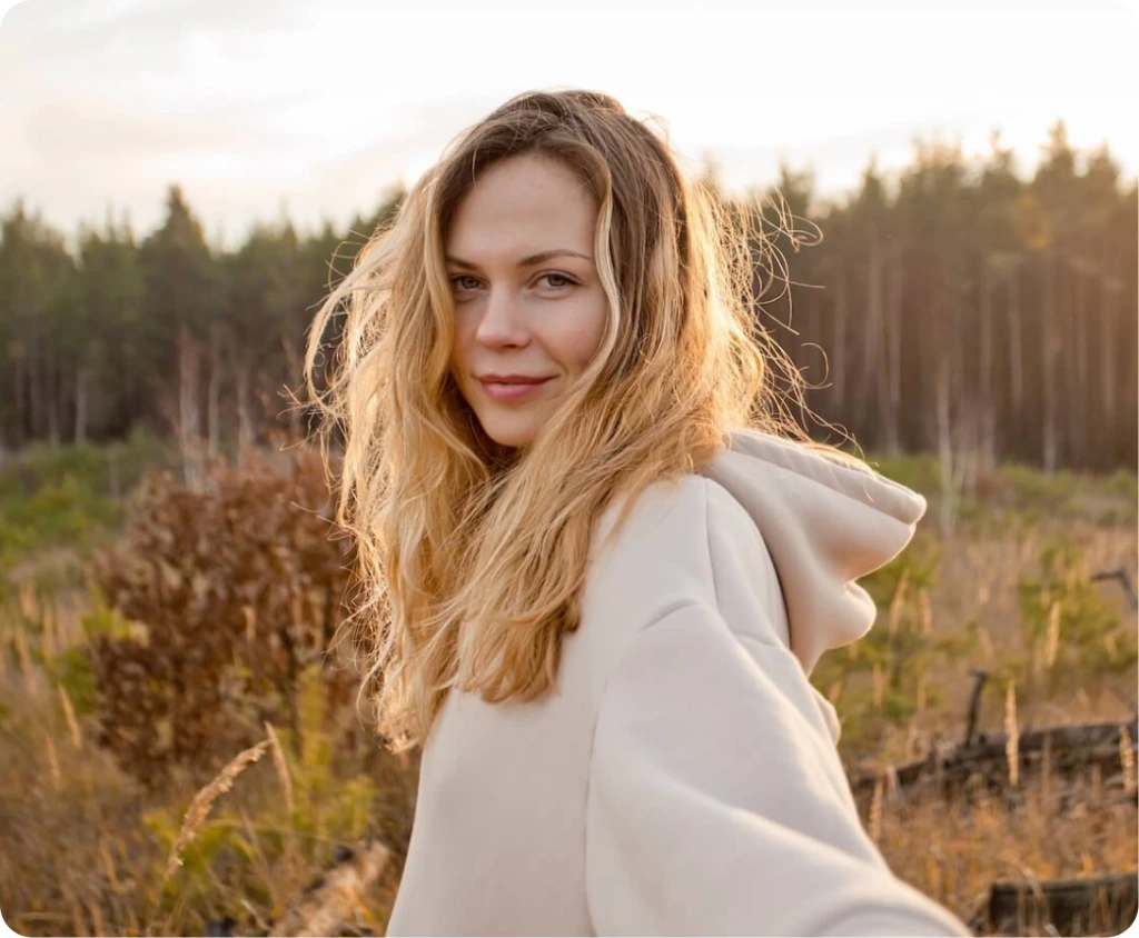 Woman with long blonde hair in a cream hoodie standing in a harvested field with evergreen trees in the background