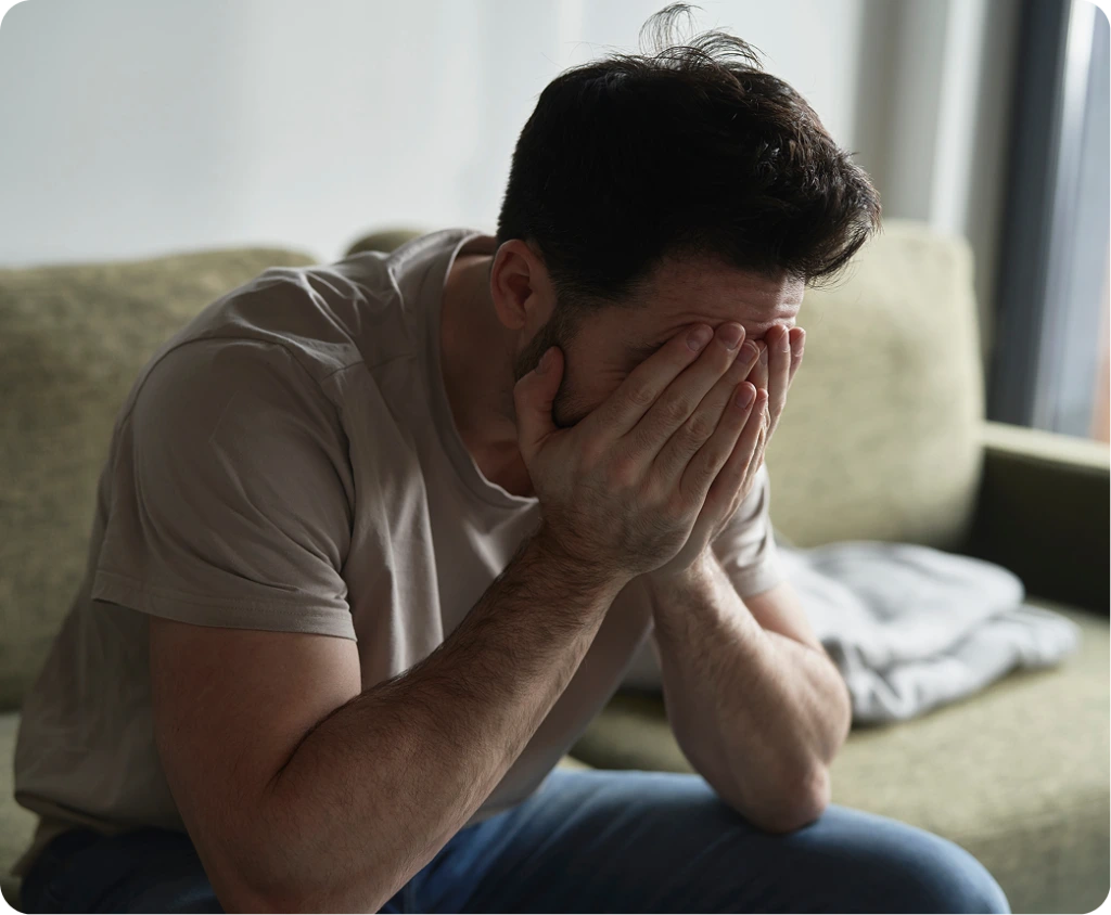 Man sitting on a couch with hands covering his face, expressing discomfort or pain