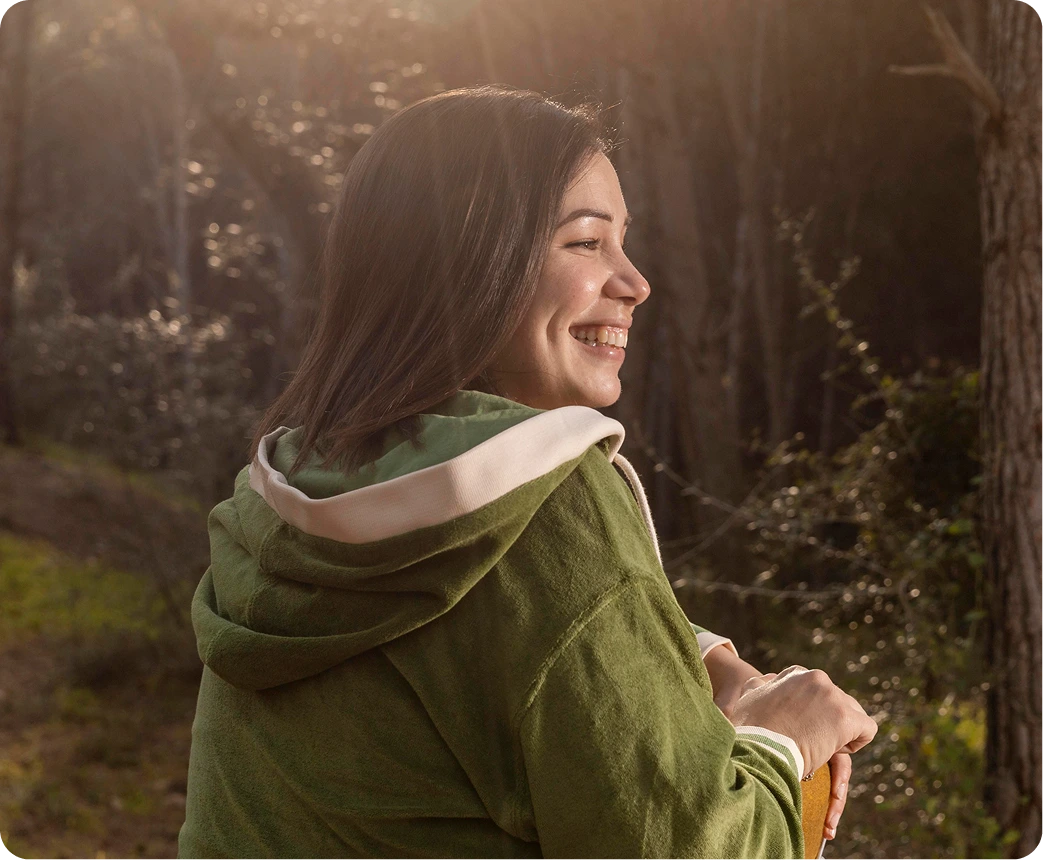 Woman in green hoodie smiling while walking through a sunlit forest