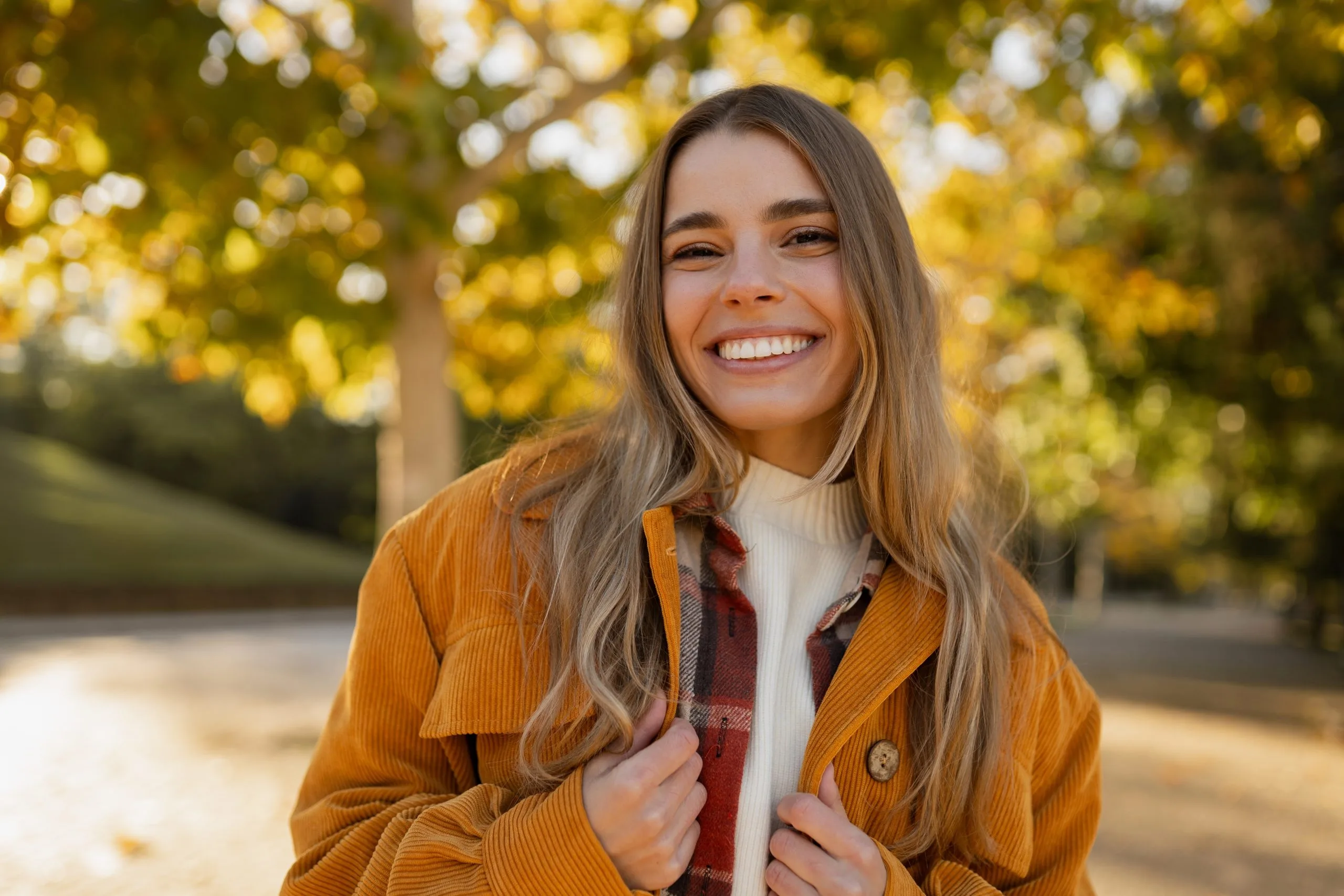 Smiling woman in a mustard jacket on a tree-lined path in autumn